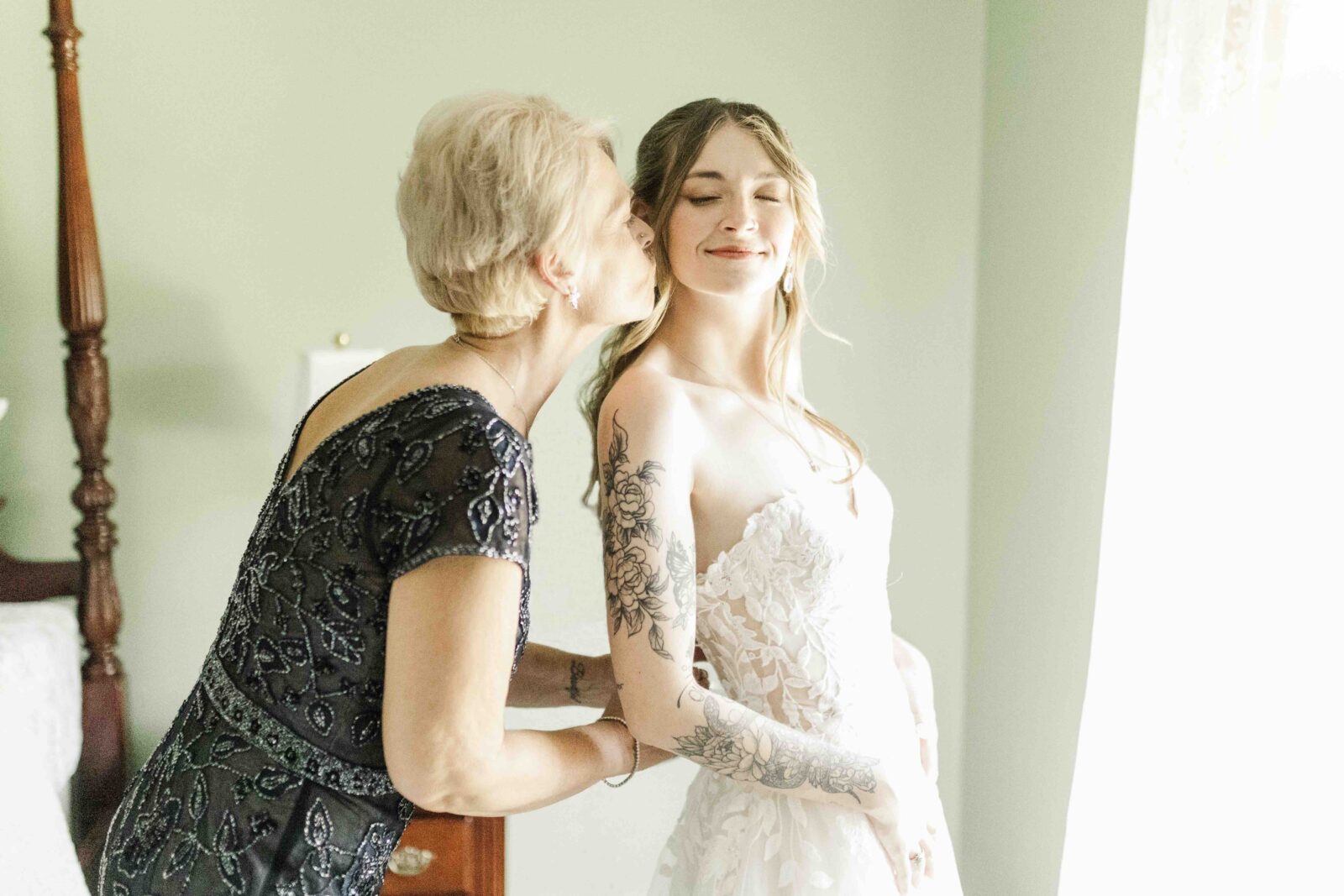 Woman kisses bride on the cheek as the bride smiles with eyes closed, standing in a softly lit room with light green walls.