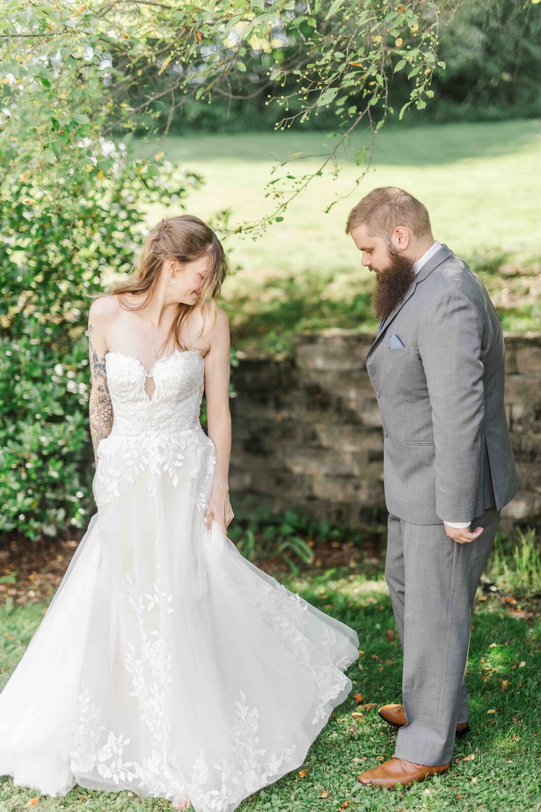 A bride in a white lace wedding dress stands on grass next to a groom in a gray suit, both looking down at the bride’s dress outside in a garden setting.