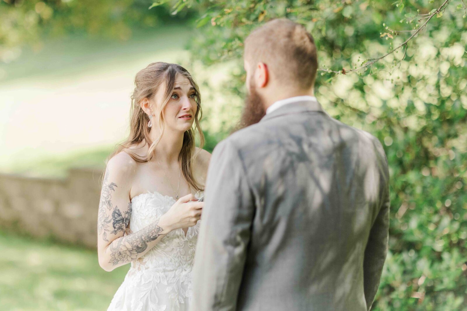 A bride in a white dress holds a piece of paper and faces a groom in a gray suit outdoors, surrounded by greenery.