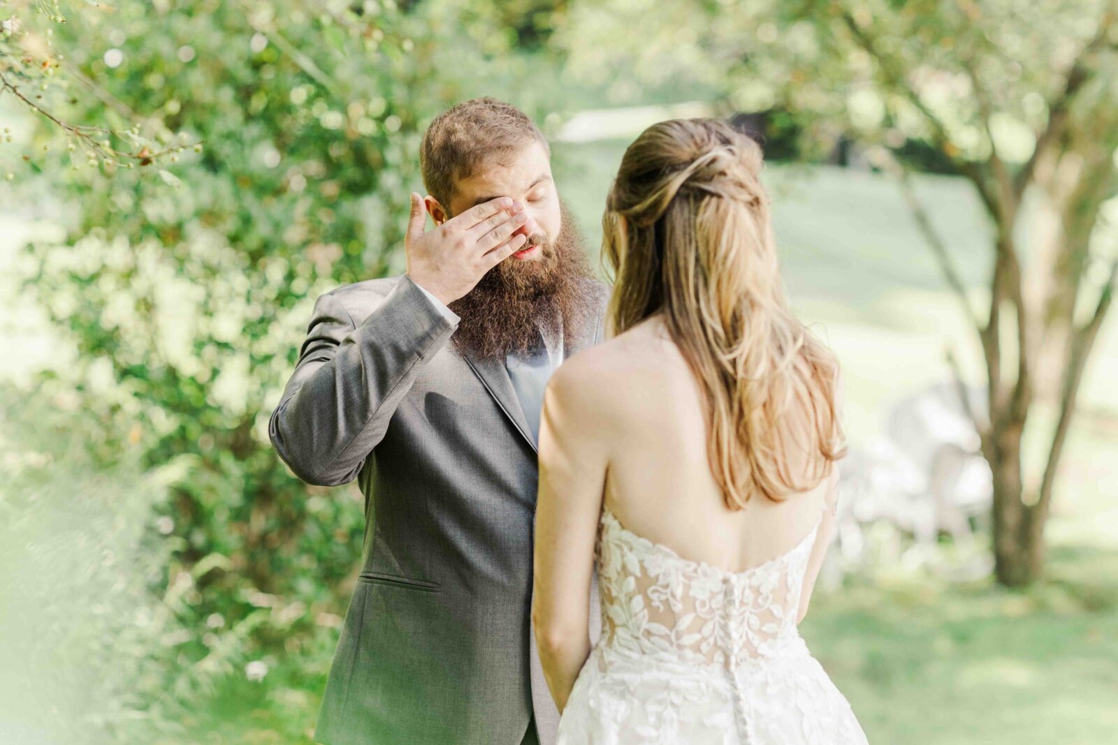 A man in a suit wipes his eyes while facing a woman in a strapless, lace wedding dress outdoors surrounded by greenery.