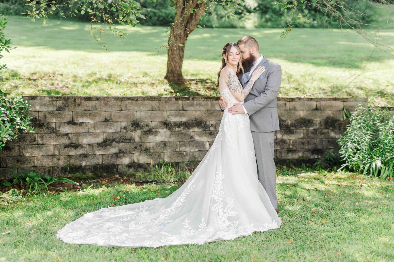 A bride in a white lace gown stands with a groom in a gray suit on grass, embracing in front of a stone wall and greenery.