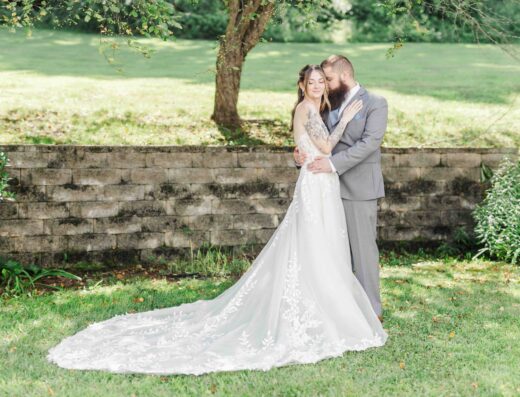 A bride in a white lace gown stands with a groom in a gray suit on grass, embracing in front of a stone wall and greenery.