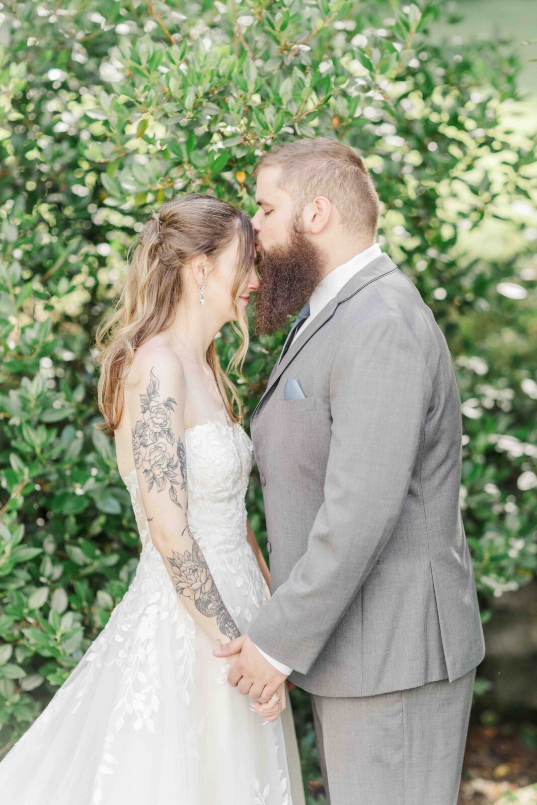 A bride and groom stand face to face, holding hands in front of green foliage. The bride wears a white dress and the groom wears a gray suit.