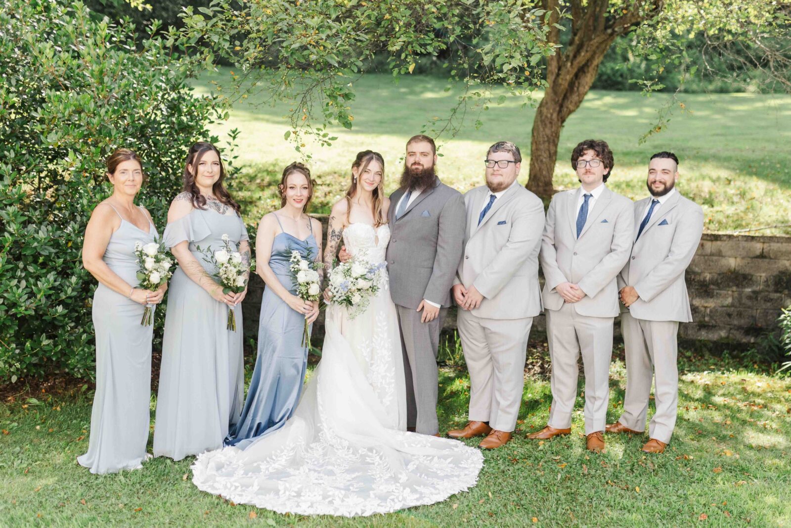 Seven people pose outdoors for a wedding photo, with four women in blue and gray dresses and three men in light suits, standing on grass in front of a tree and greenery.