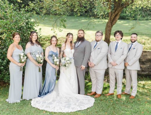Seven people pose outdoors for a wedding photo, with four women in blue and gray dresses and three men in light suits, standing on grass in front of a tree and greenery.