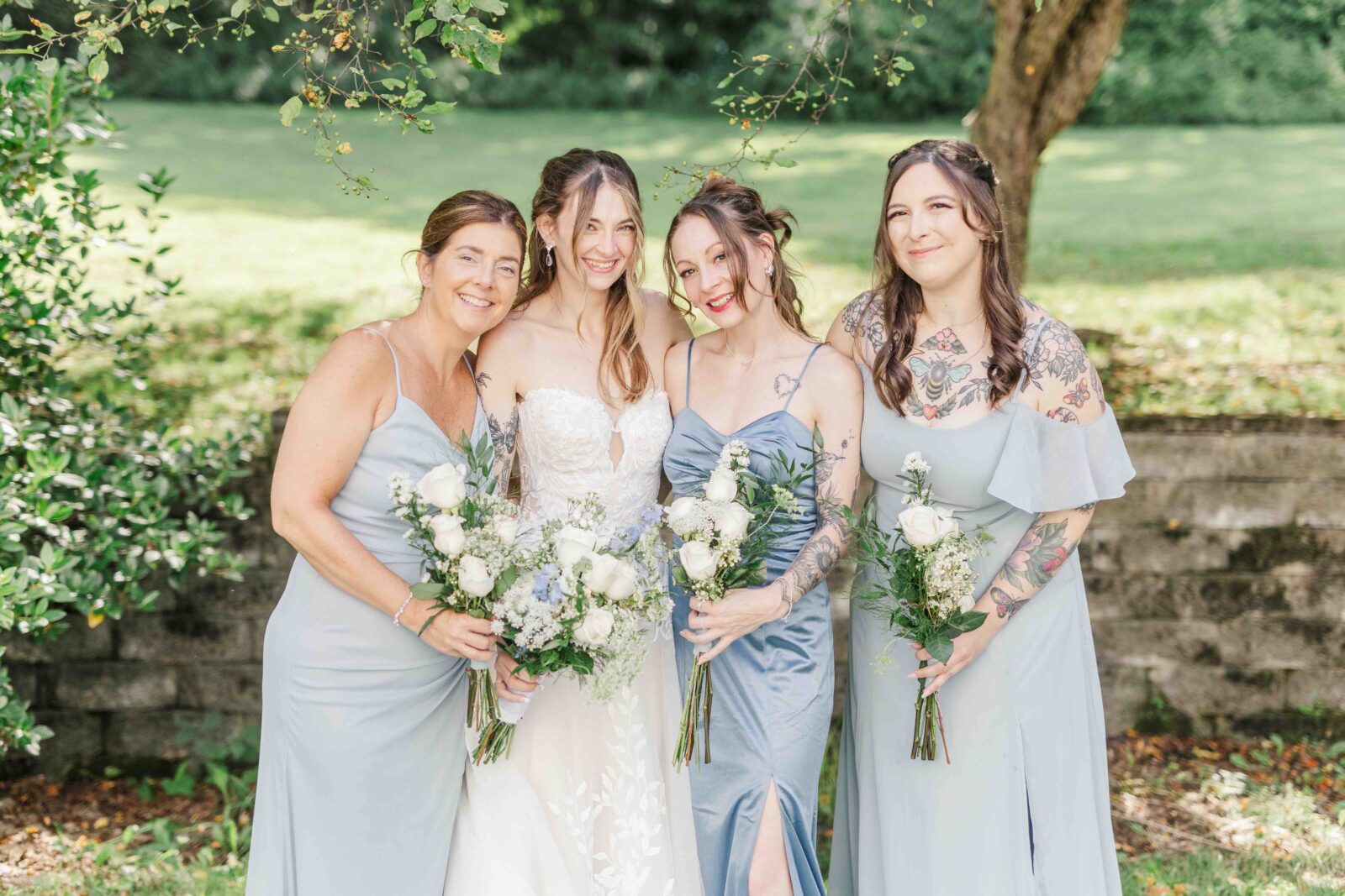 Bride in white with three bridesmaids in dusty light blue dresses holding white rose and greenery bouquets outdoors under trees at a backyard summer wedding