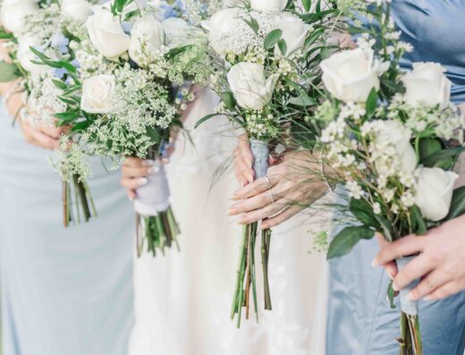 Three women in light-colored dresses hold bouquets of white roses and greenery, standing side by side outdoors.