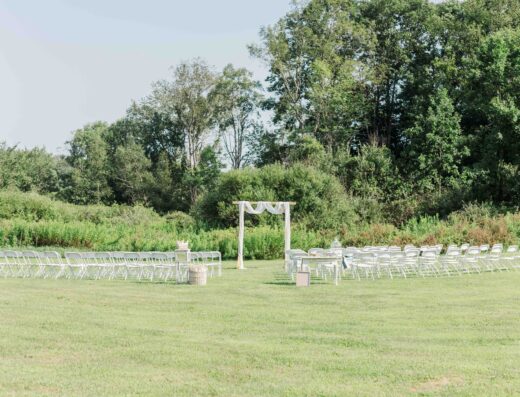 Outdoor wedding ceremony setup with white chairs arranged in rows facing a simple wooden arch draped with white fabric on a grassy field, surrounded by trees and greenery.