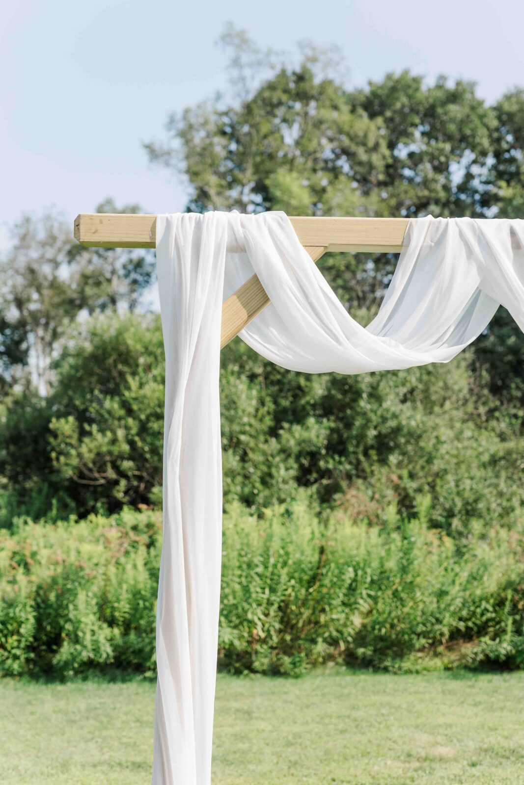 A wooden arch draped with white fabric stands outdoors in front of green trees and grass under a clear sky.
