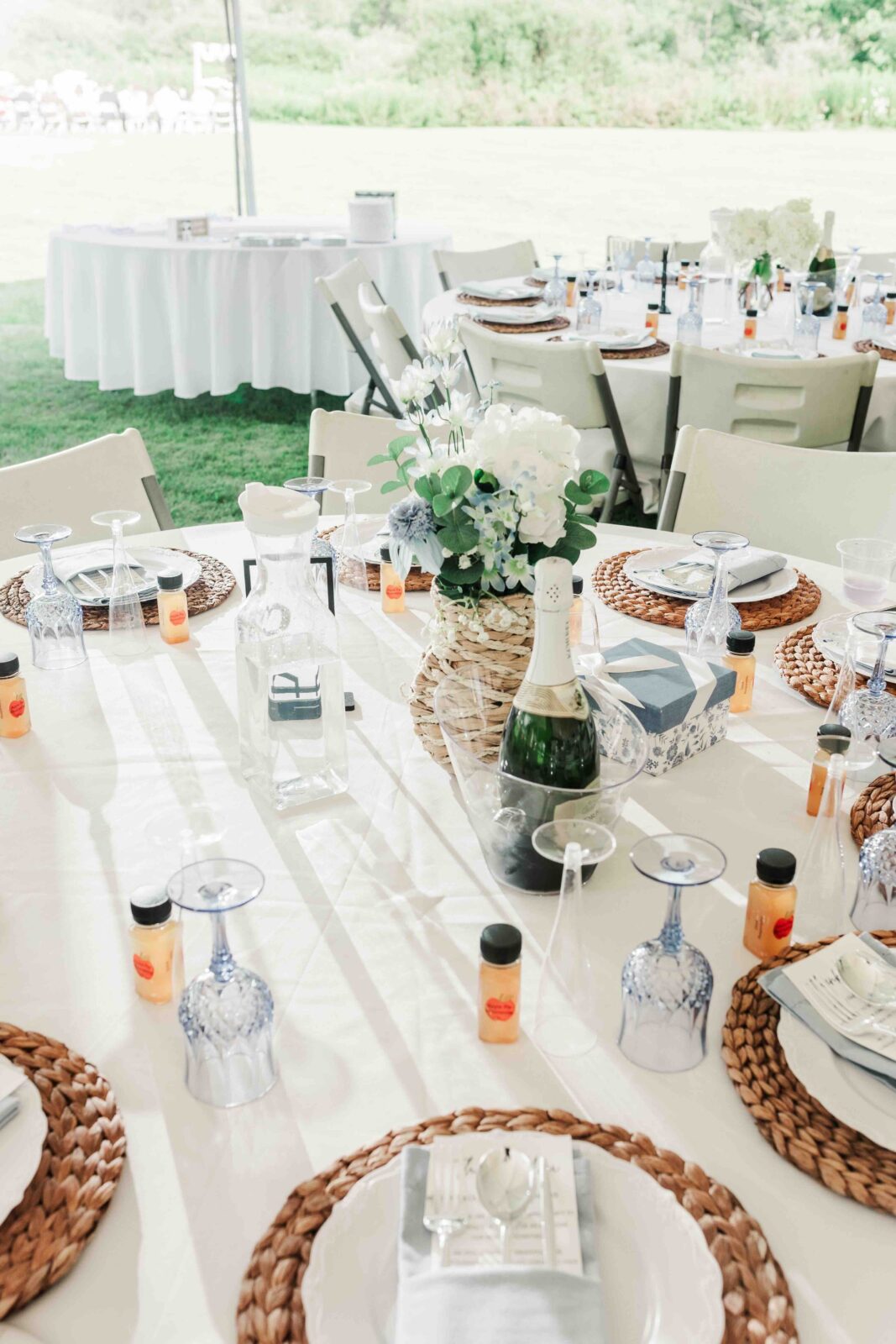 Outdoor event table set with woven placemats, plates, blue glasses, mini orange bottles, lanterns, a champagne bottle, and a white flower centerpiece on a white tablecloth.