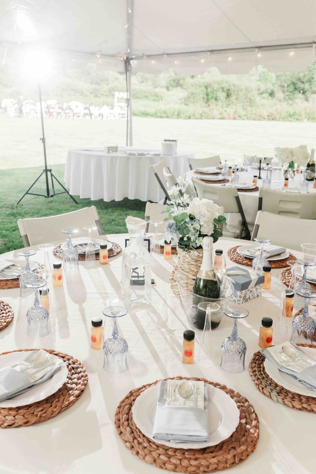 Outdoor event table set with woven placemats, glassware, champagne, small bottles, white napkins, and a floral centerpiece under a tent.