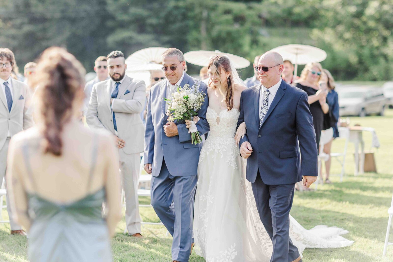 A bride in a white dress walks down the aisle outdoors, escorted by two men in suits. Guests watch in the background on a sunny day.