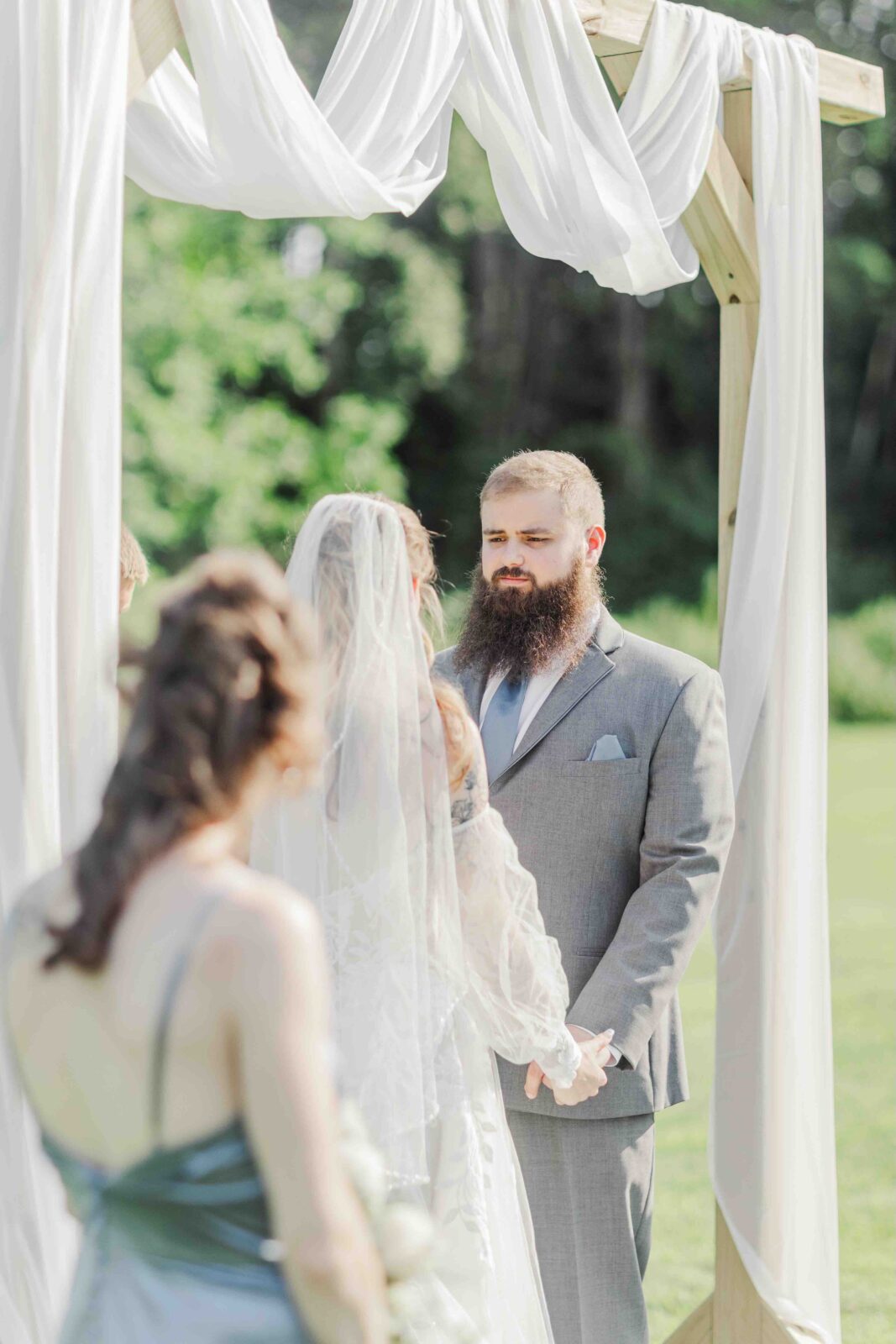 A bride and groom stand holding hands under a wooden arch draped with white fabric during an outdoor wedding ceremony.