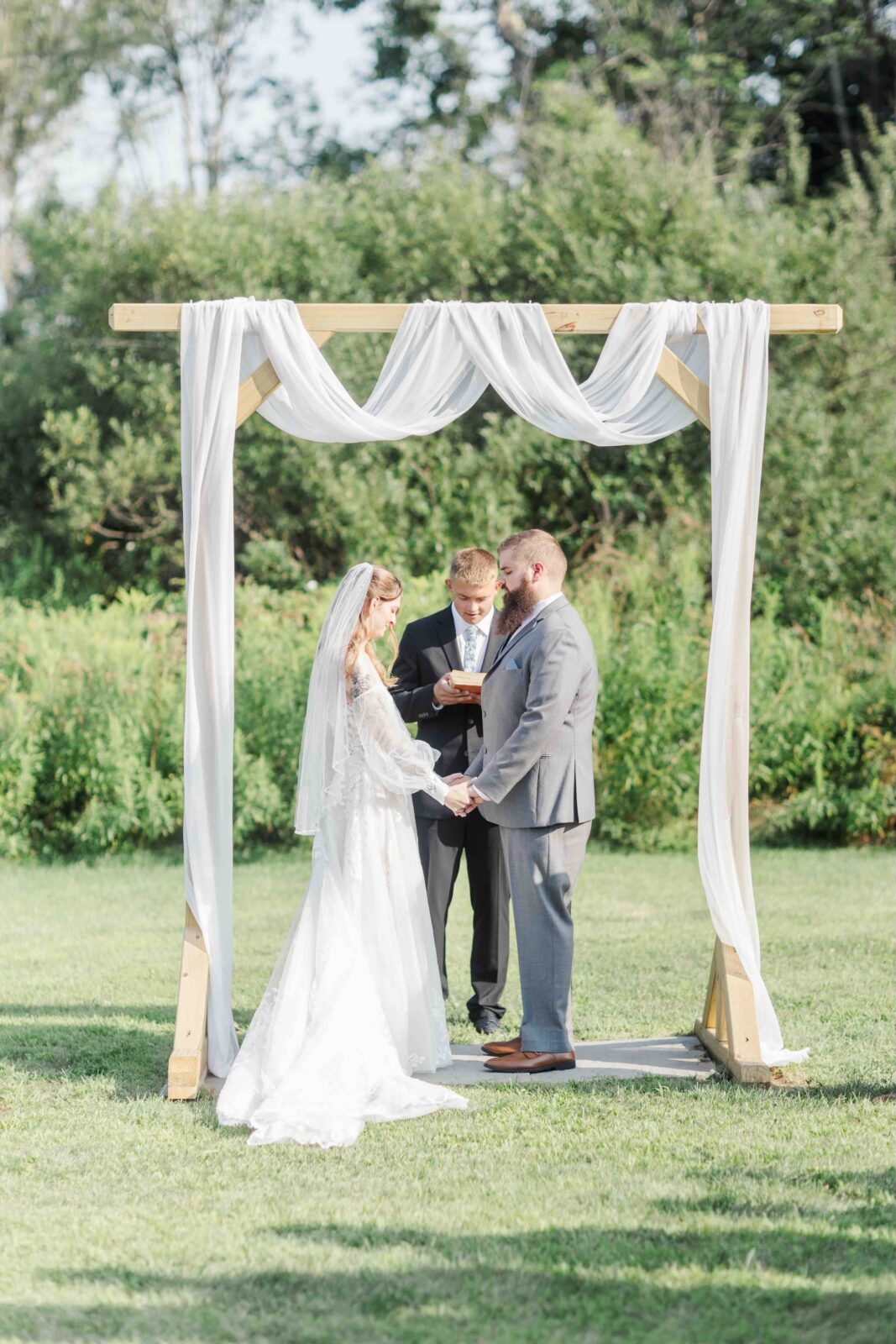 A bride and groom stand under a wooden arch draped with white fabric, holding hands during an outdoor wedding ceremony as an officiant stands behind them.