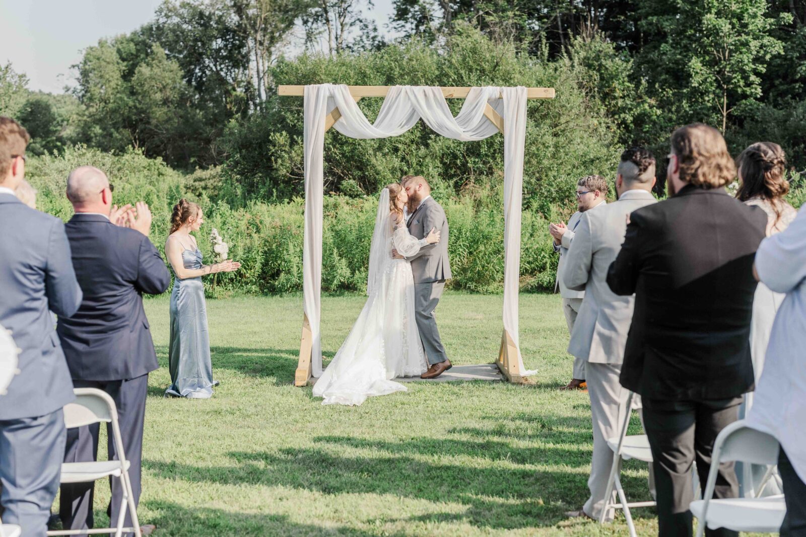 A bride and groom kiss under a decorated arch outdoors as wedding guests seated on chairs watch and applaud.