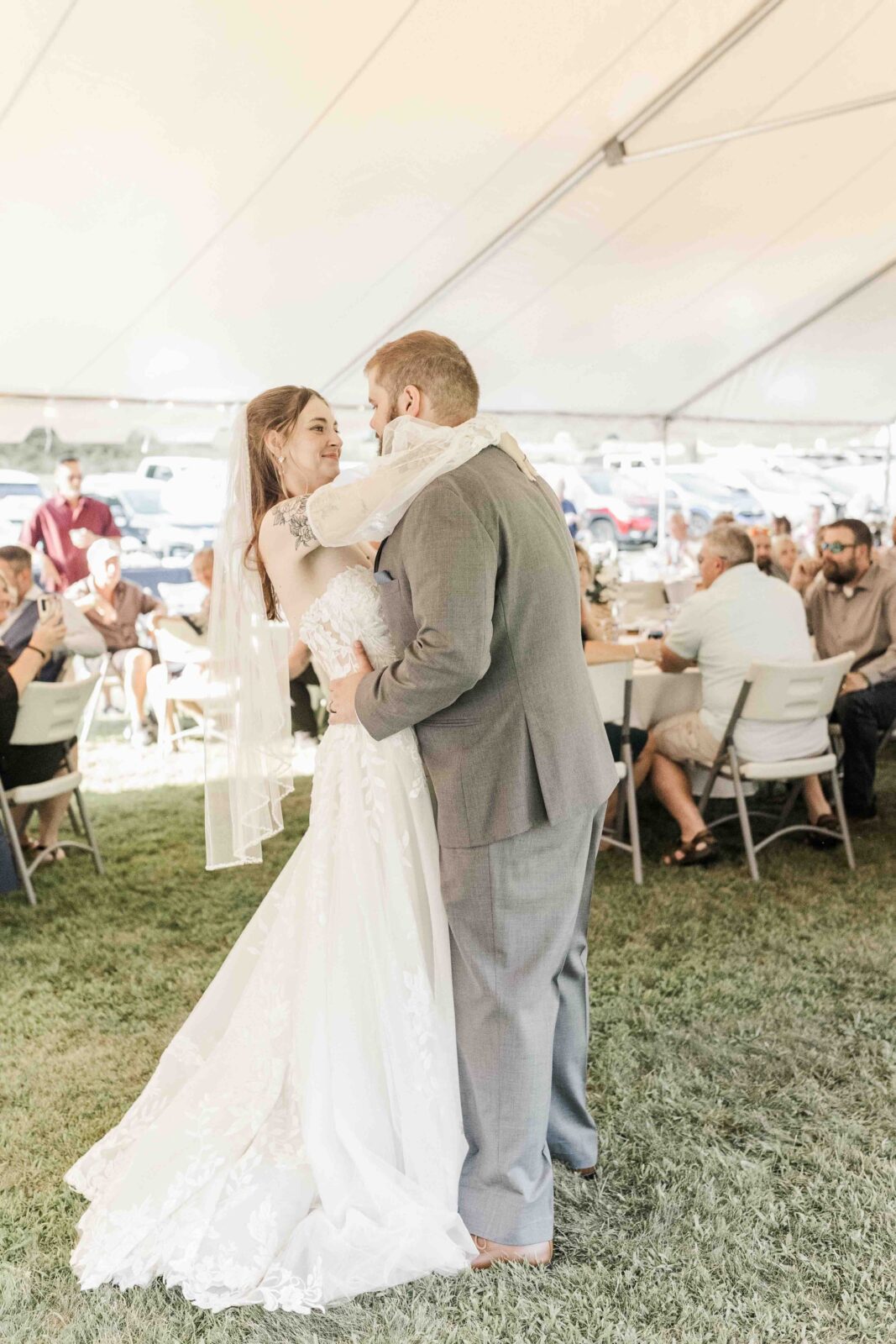A bride and groom share their first dance under a white tent at an outdoor wedding reception, with guests seated at tables in the background.