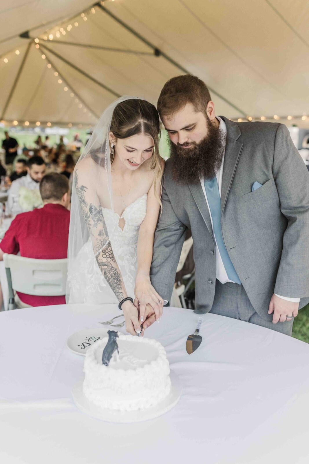 Bride and groom in wedding attire cut a white cake together under a tent, with guests seated in the background.