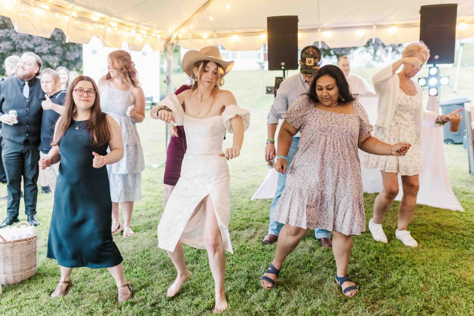 A group of people dance together on grass under a tent with string lights. A woman in a white dress and hat stands in front, leading the group.
