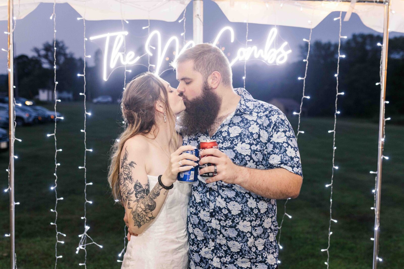 A couple shares a kiss while holding cans, standing in front of string lights and a neon sign that reads “The McCombs” at an outdoor event.