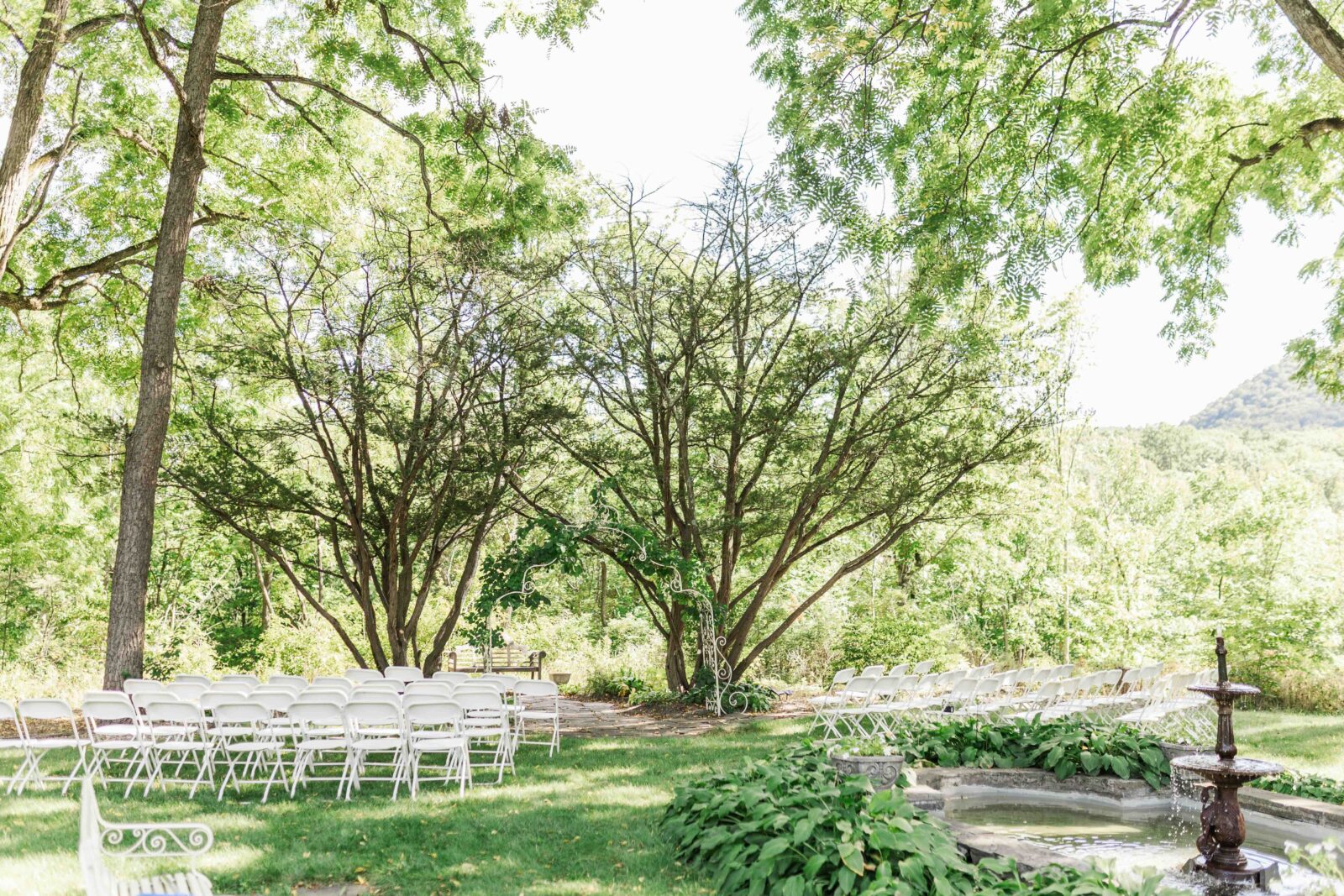 Rows of white folding chairs arranged outdoors on grass under leafy trees, facing a small arbor, with a fountain visible to the right.