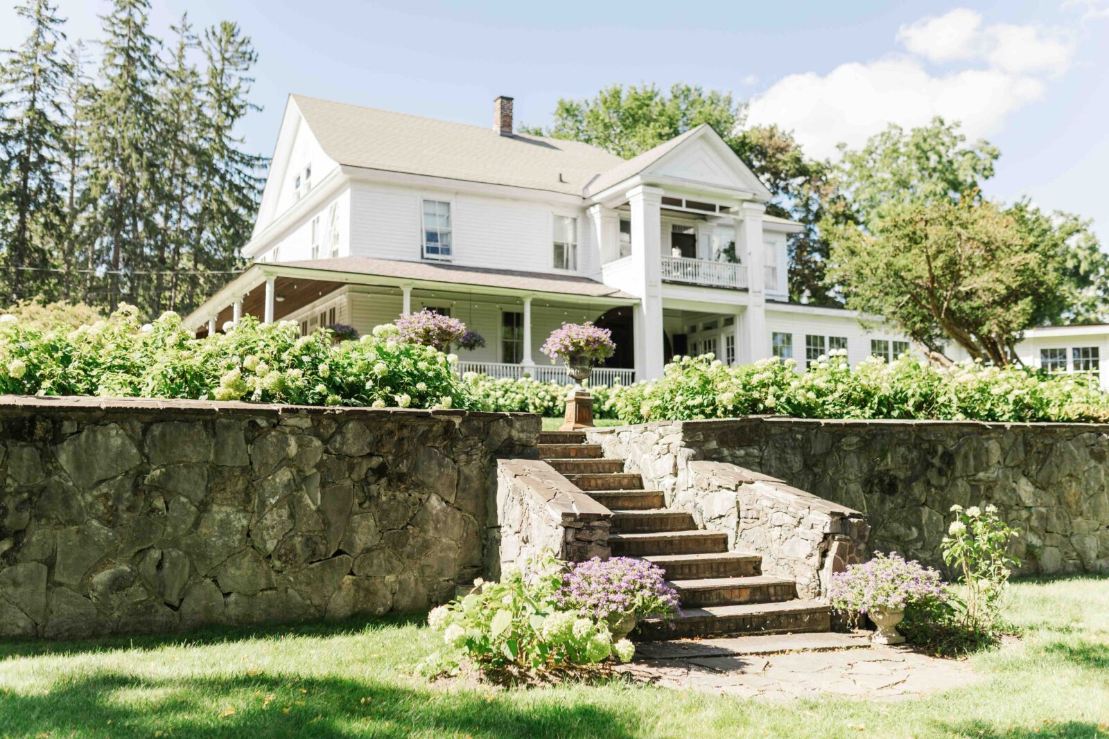 A large white house with a wraparound porch sits behind a stone wall, with steps leading up through a garden filled with blooming flowers and greenery.