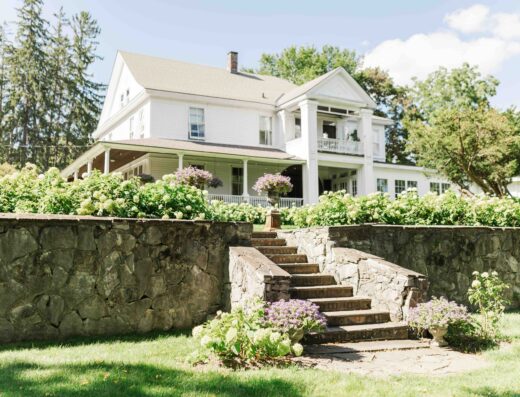 A large white house with a wraparound porch sits behind a stone wall, with steps leading up through a garden filled with blooming flowers and greenery.