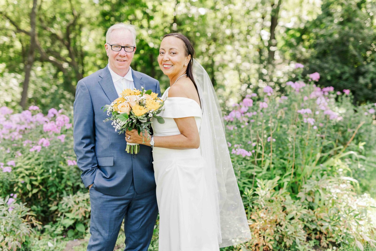 A couple poses outdoors, with the woman in a white wedding dress holding a bouquet and the man in a blue suit, surrounded by green foliage and purple flowers.