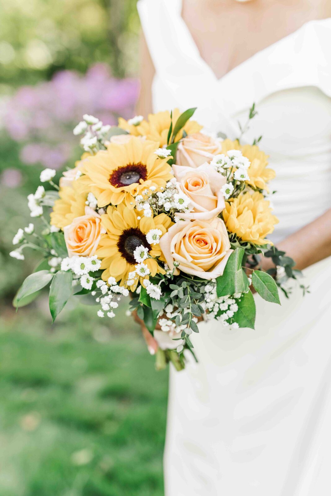 A person in a white dress holds a bouquet of sunflowers, pale pink roses, baby’s breath, and greenery outdoors.