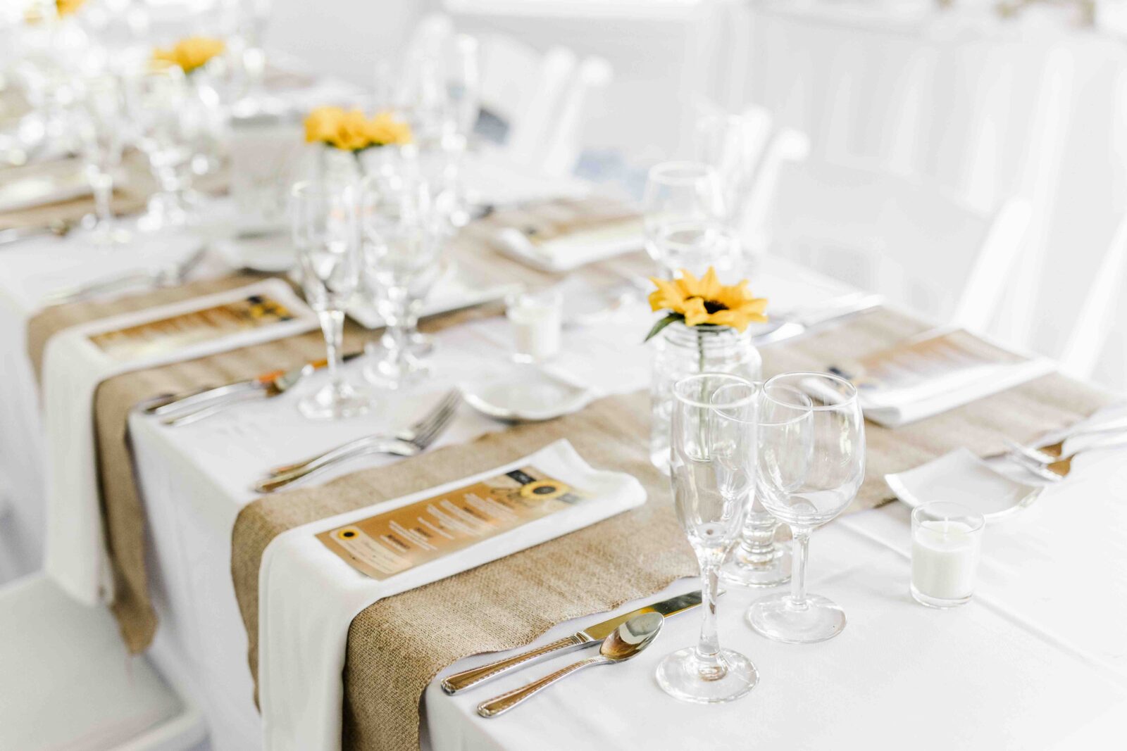 A formal table setting with white linens, burlap runners, menus, glassware, utensils, and small sunflower centerpieces on each table.