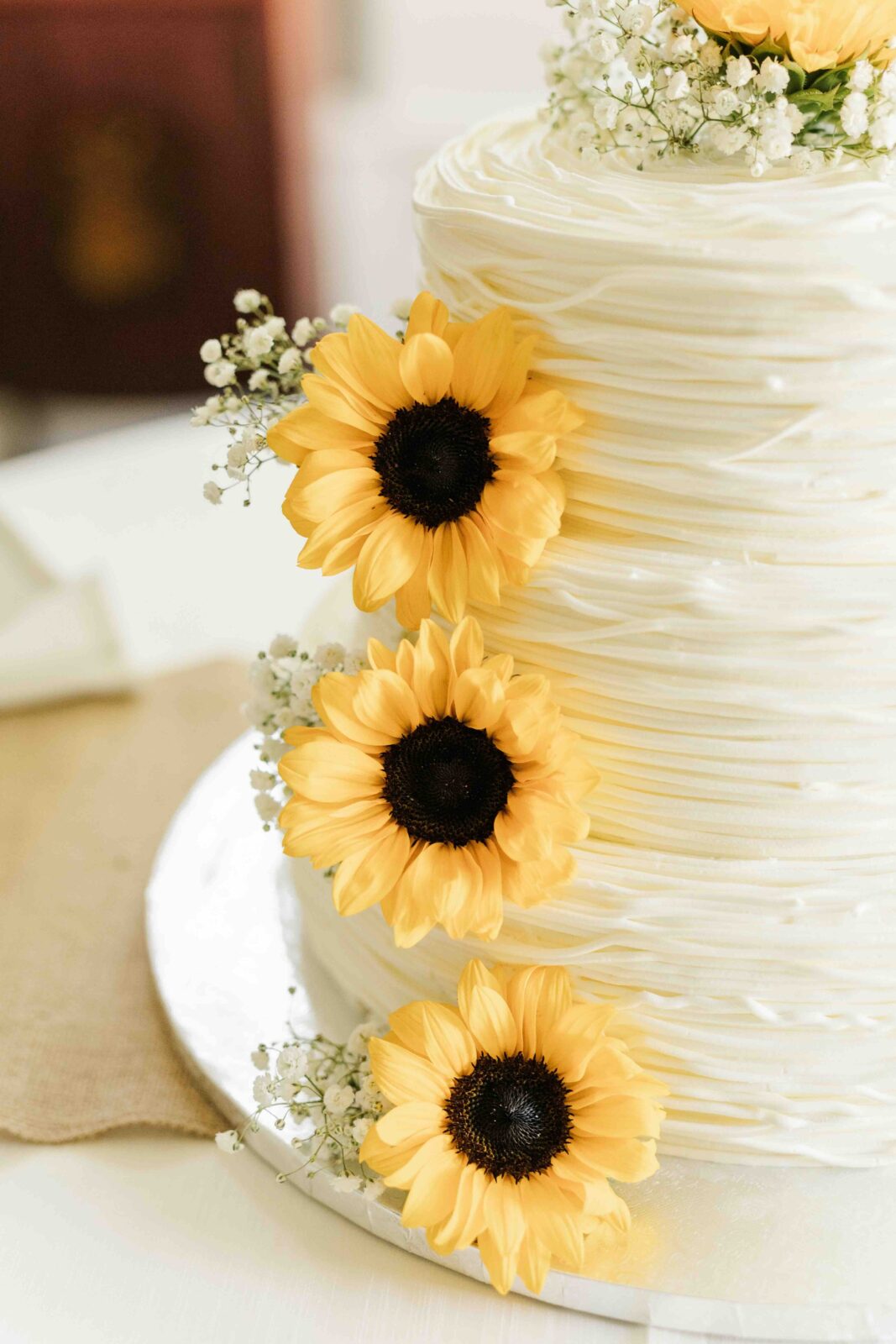 Close-up of a white, textured tiered cake decorated with three yellow sunflowers and small white flowers on the side.