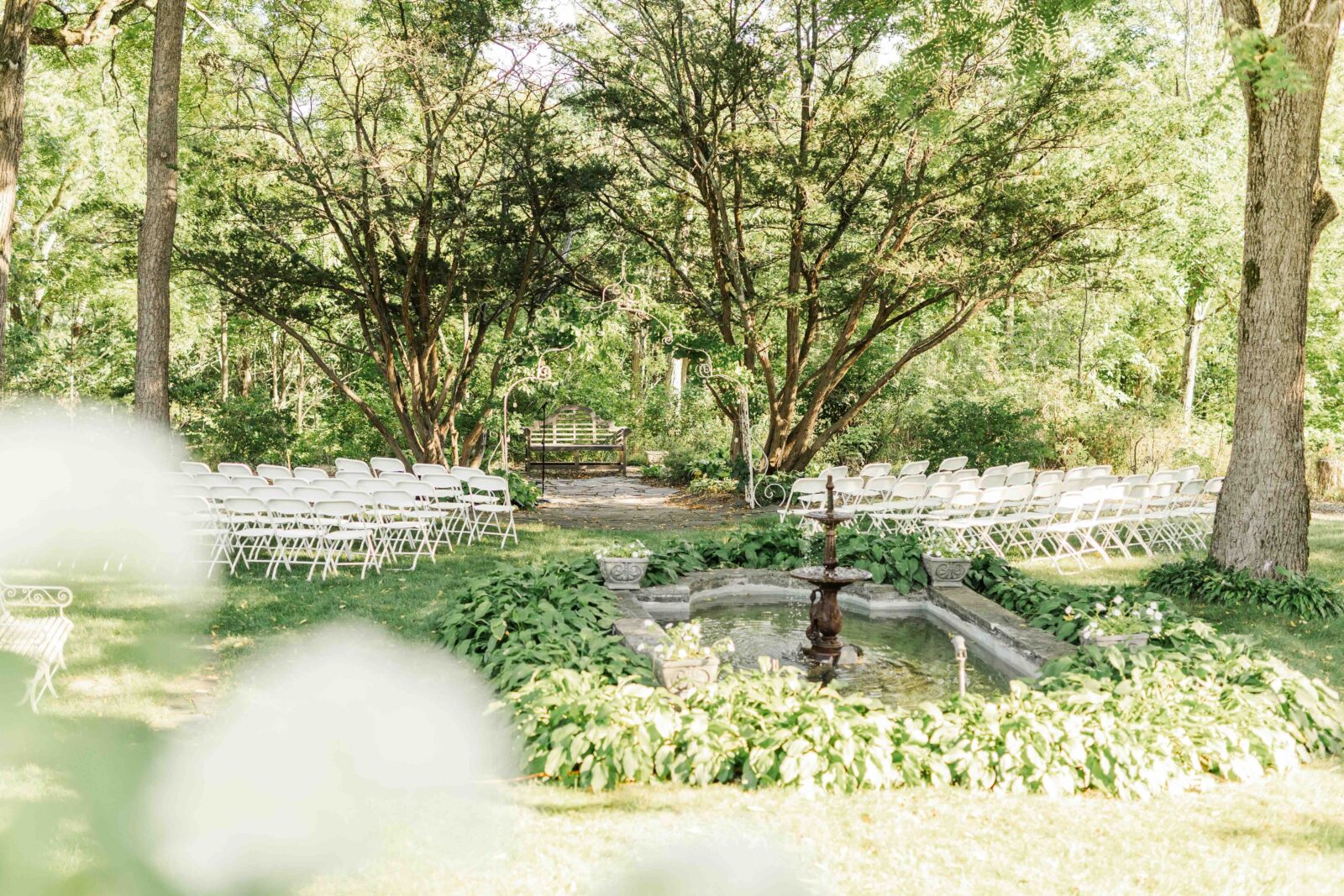 Rows of white chairs face a small fountain in a garden, surrounded by trees and greenery, set up for an outdoor event.