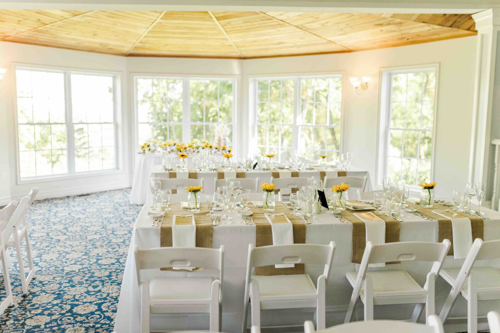 Bright banquet room with large windows, white chairs, and tables set with glassware, plates, and small yellow flower centerpieces on beige runners.