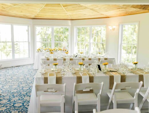 Bright banquet room with large windows, white chairs, and tables set with glassware, plates, and small yellow flower centerpieces on beige runners.