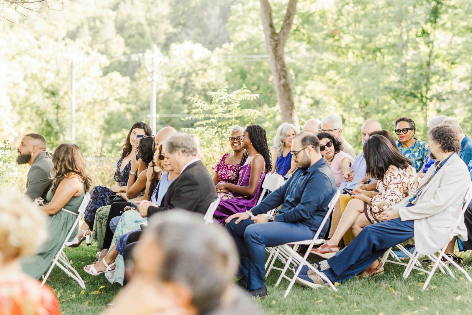 A group of people sit on white folding chairs outdoors, surrounded by greenery, attending a formal event in daylight.