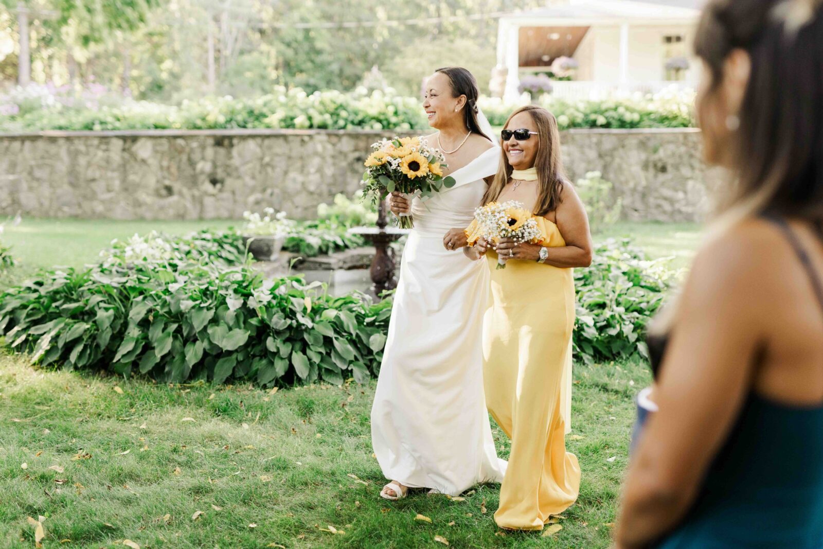 Two women in yellow dresses, one holding a bouquet of sunflowers, walk together outdoors on grass, smiling, with greenery and a stone wall in the background.