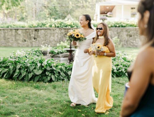 Two women in yellow dresses, one holding a bouquet of sunflowers, walk together outdoors on grass, smiling, with greenery and a stone wall in the background.