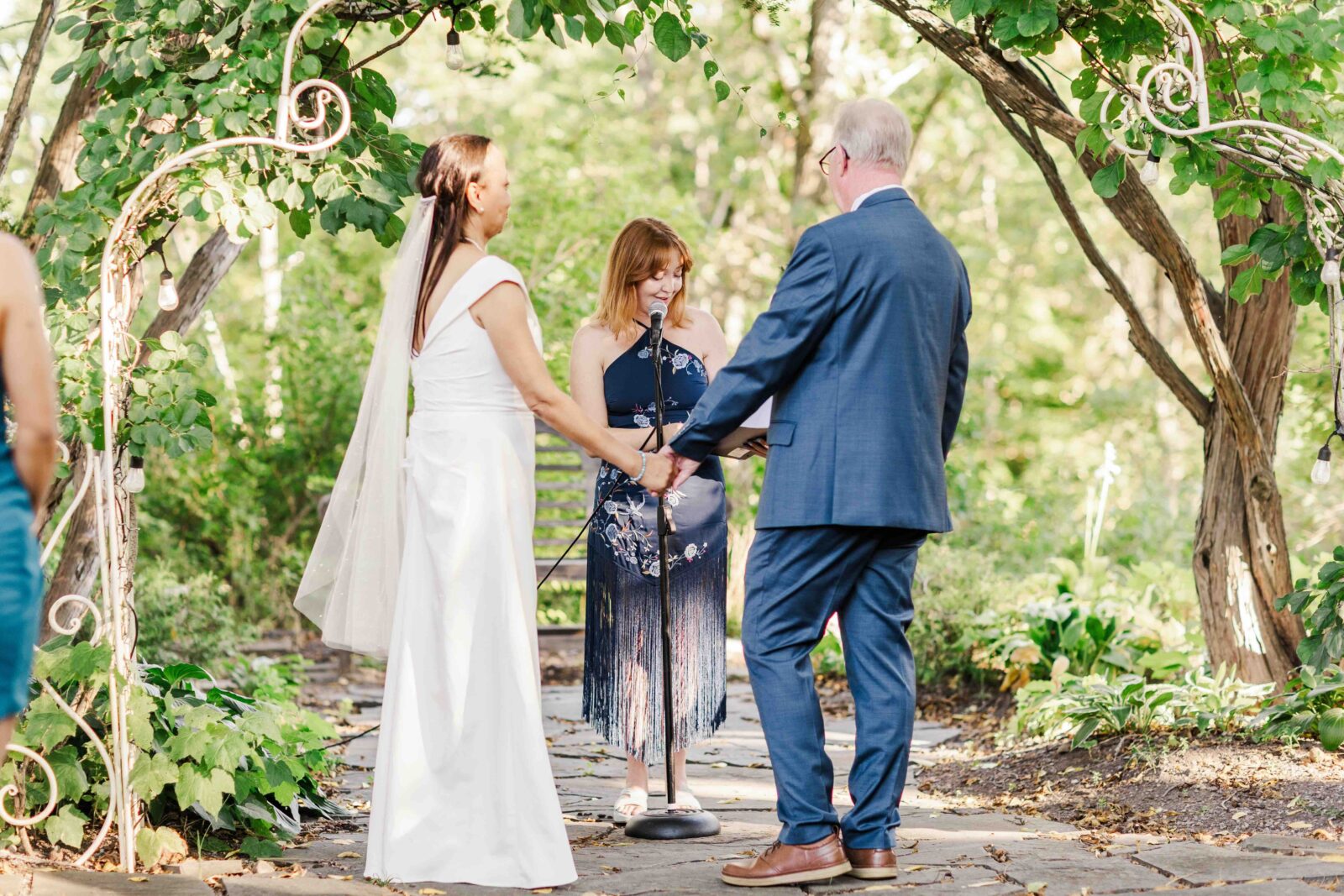 A bride and groom hold hands during an outdoor wedding ceremony, with an officiant standing in front of them under a leafy arch.