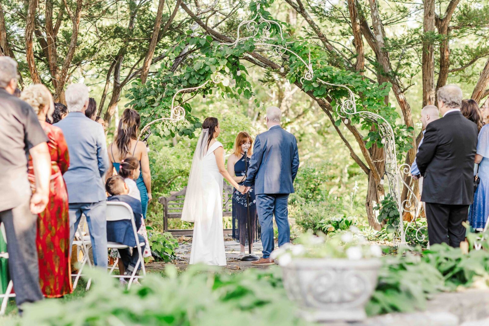 A couple holding hands during an outdoor wedding ceremony under a floral archway, with a wedding officiant standing between them and guests watching from behind.