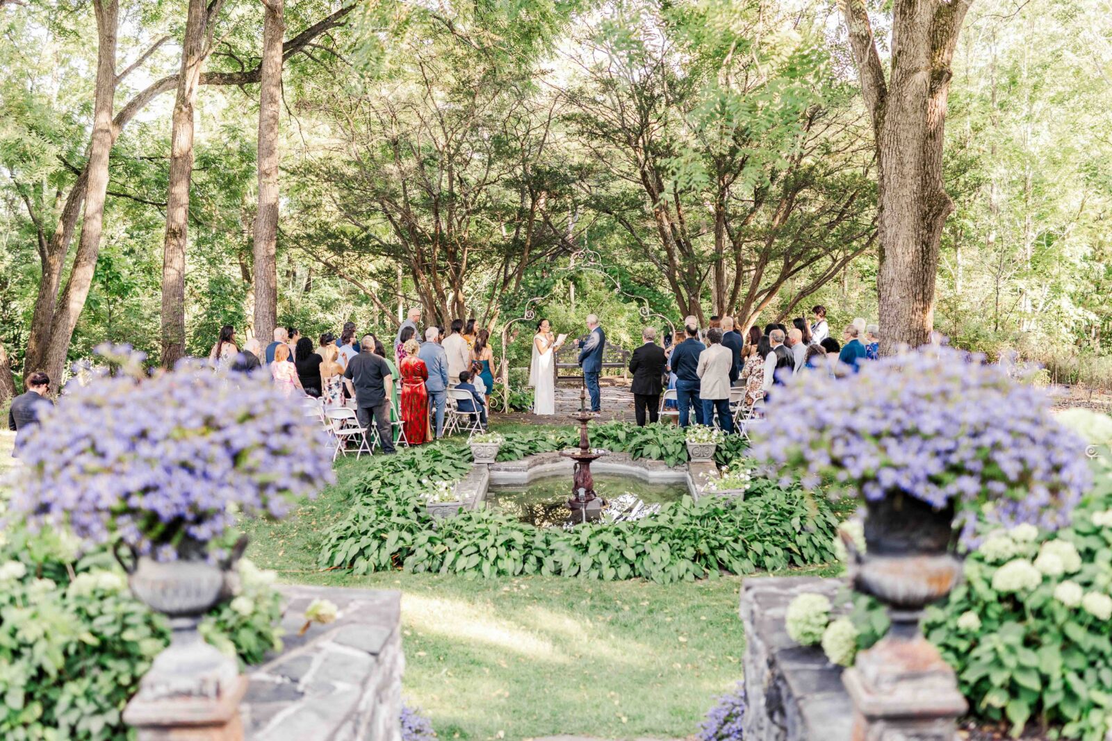 An outdoor wedding ceremony is taking place in a garden, with guests seated and a couple standing at the altar under tall trees.