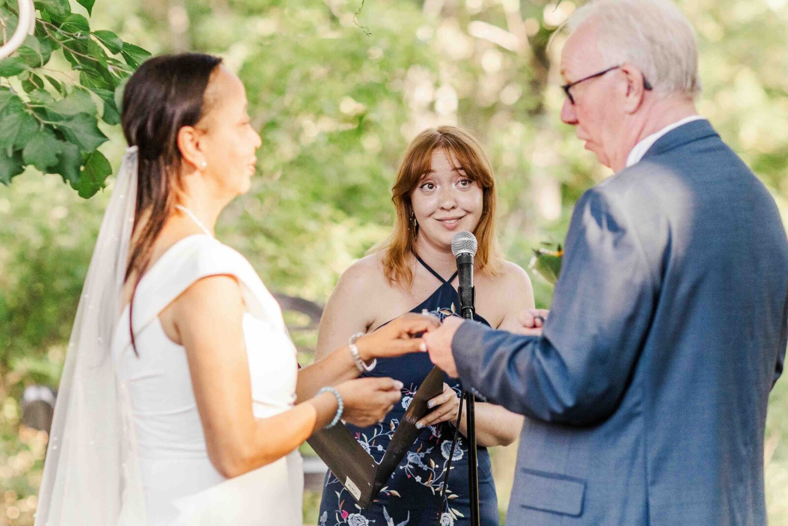 A woman and man exchange rings during an outdoor wedding ceremony as an officiant stands behind a microphone, watching them.