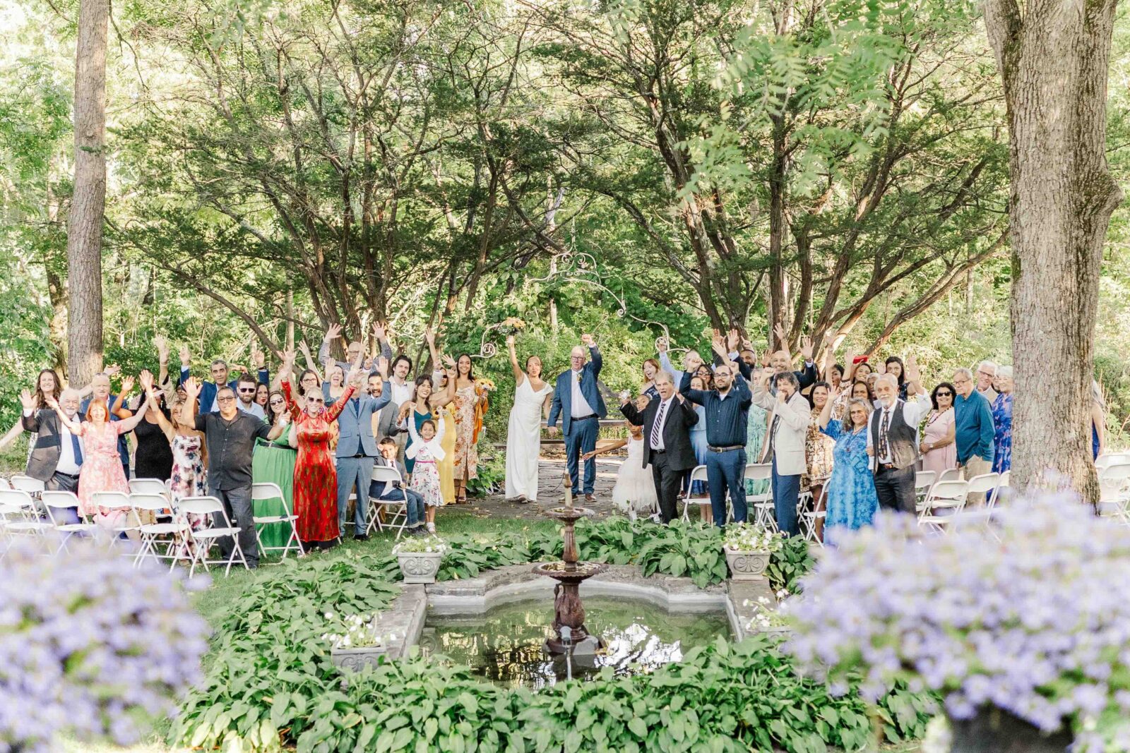 A large group of people stands and waves near a small outdoor fountain during a ceremony in a wooded garden setting with white chairs.