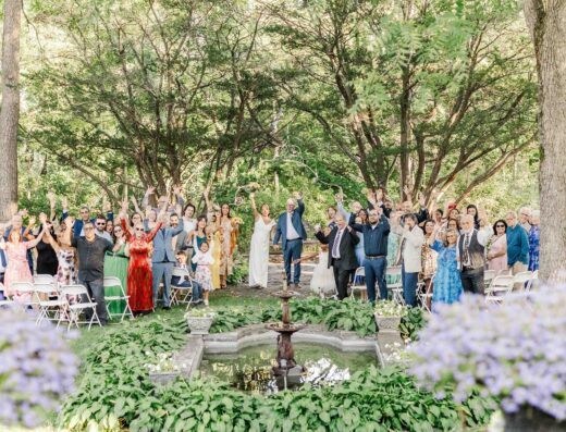 A large group of people stands and waves near a small outdoor fountain during a ceremony in a wooded garden setting with white chairs.