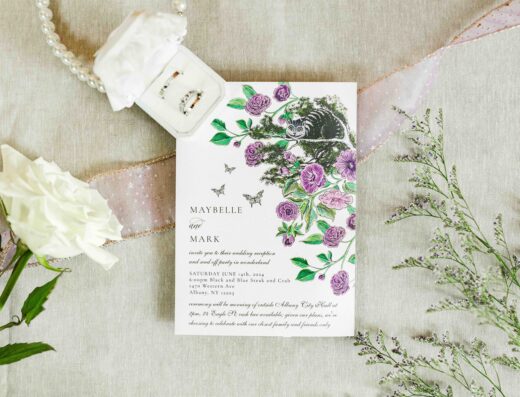 A floral wedding invitation with purple flowers lies on a beige surface, surrounded by a white rose, greenery, a ring box with rings, a pearl necklace, and a pink ribbon.