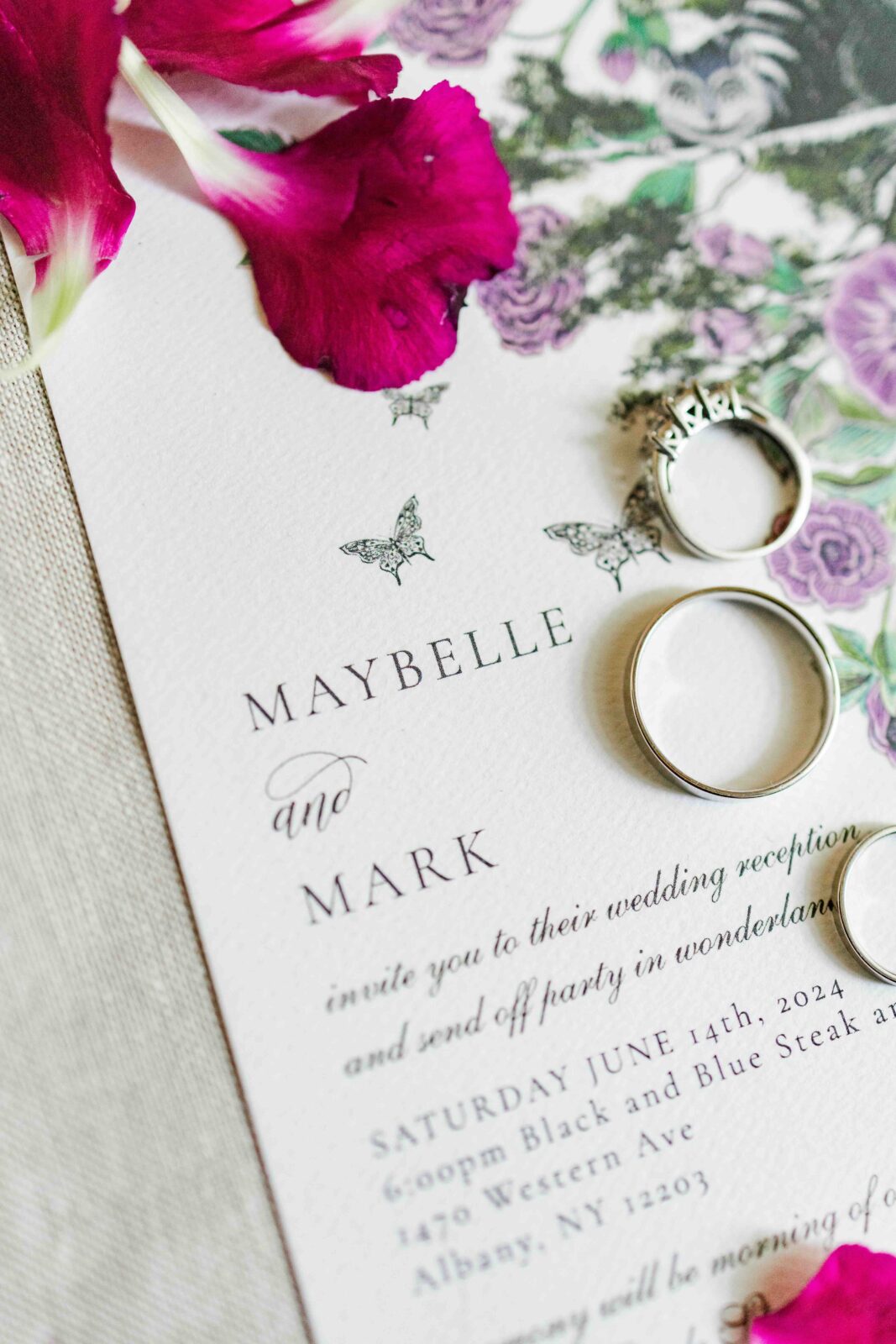 Close-up of a wedding invitation with floral design, two rings, and flower petals. The invitation is addressed to Maybelle and Mark for a June 14, 2024 wedding.