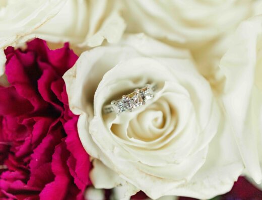 A silver diamond ring rests on a white rose, surrounded by white and deep pink flowers.