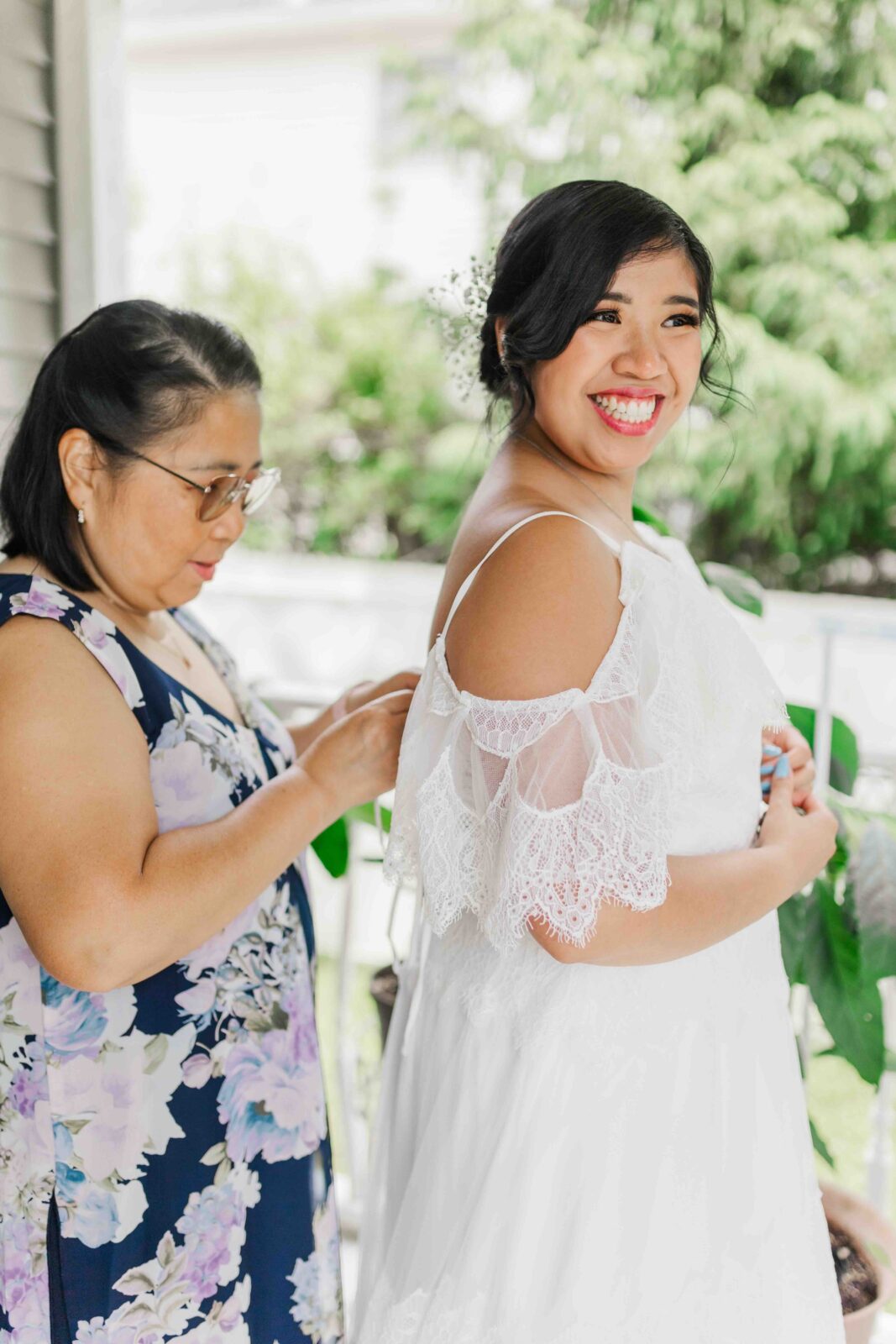A woman in a floral dress fastens the back of a bride’s white lace gown as the bride smiles and stands facing forward.