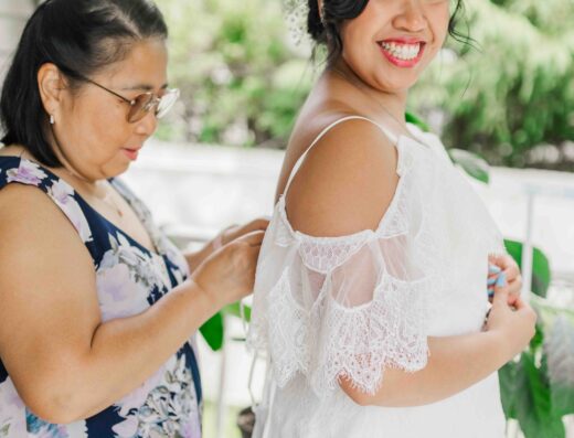 A woman in a floral dress fastens the back of a bride’s white lace gown as the bride smiles and stands facing forward.