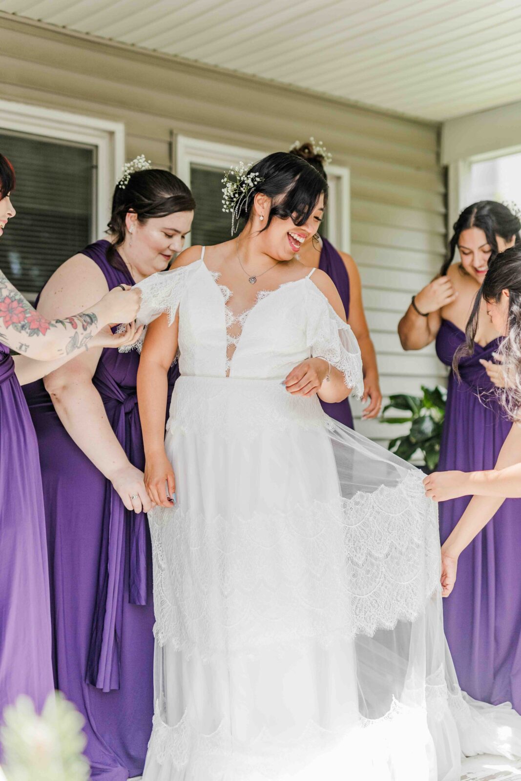 A bride in a white dress stands smiling as four bridesmaids in purple dresses help arrange her gown under a covered outdoor area.