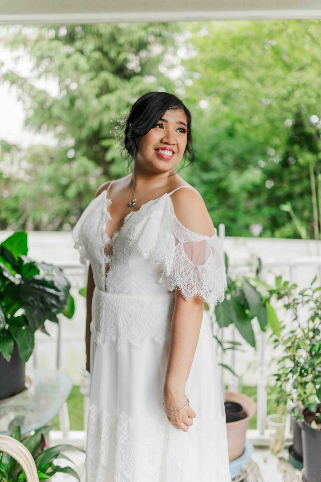 Woman in a white lace dress stands on a porch surrounded by potted plants, smiling and looking to the side.