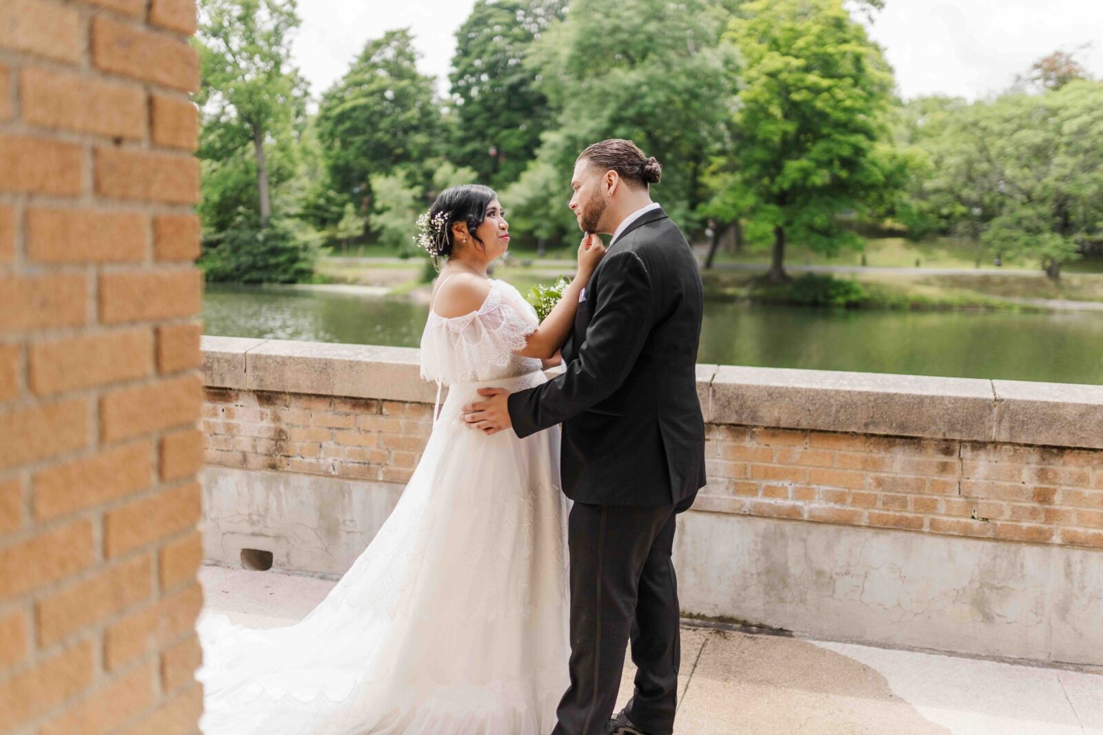 A bride and groom stand outside by a stone wall overlooking a lake and trees, gazing at each other on their wedding day.